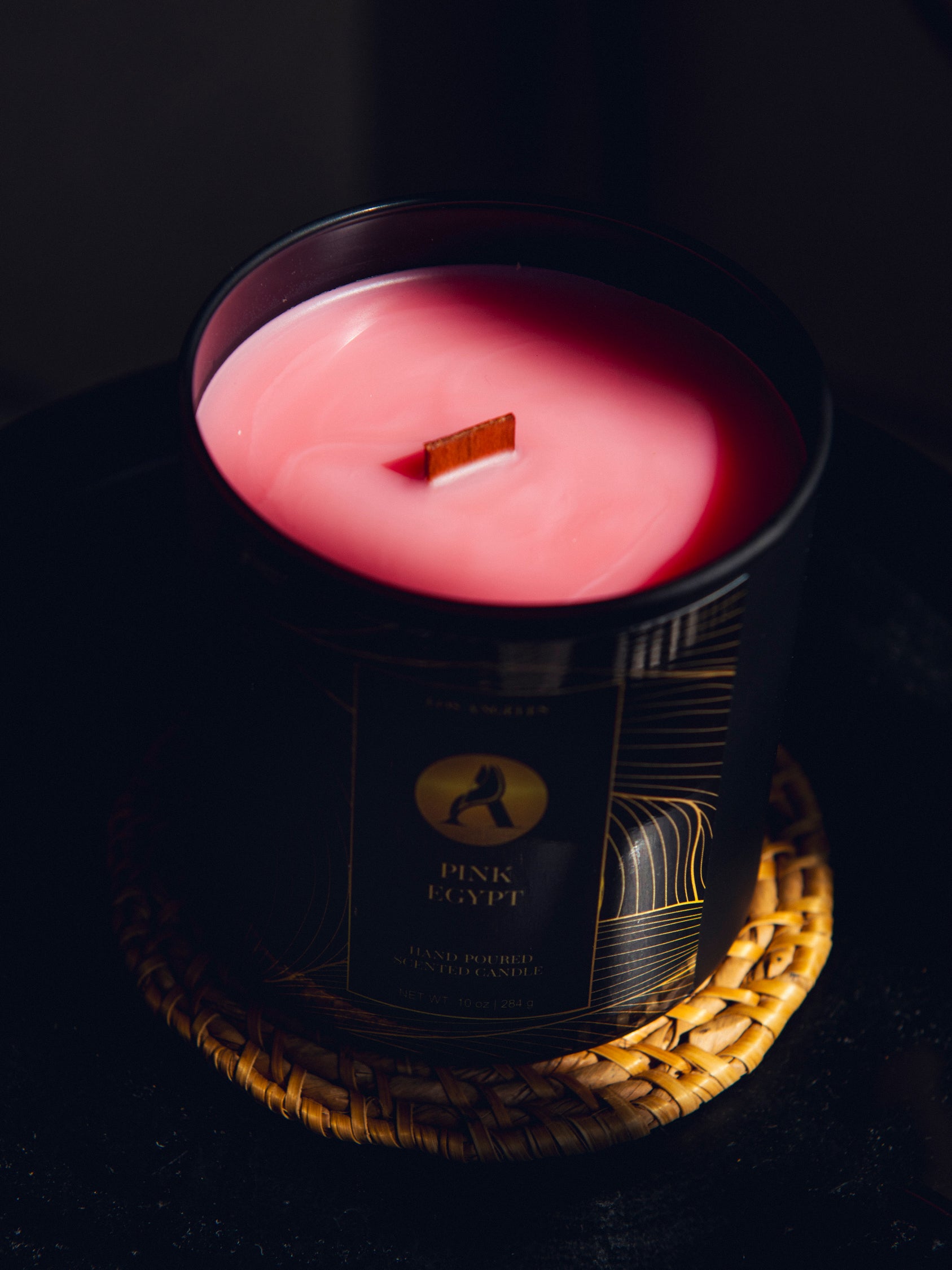 A black candle jar with pink wax and a wooden wick, resting on a woven straw coaster against a dark background. The label reads 'PINK EGYPT'.