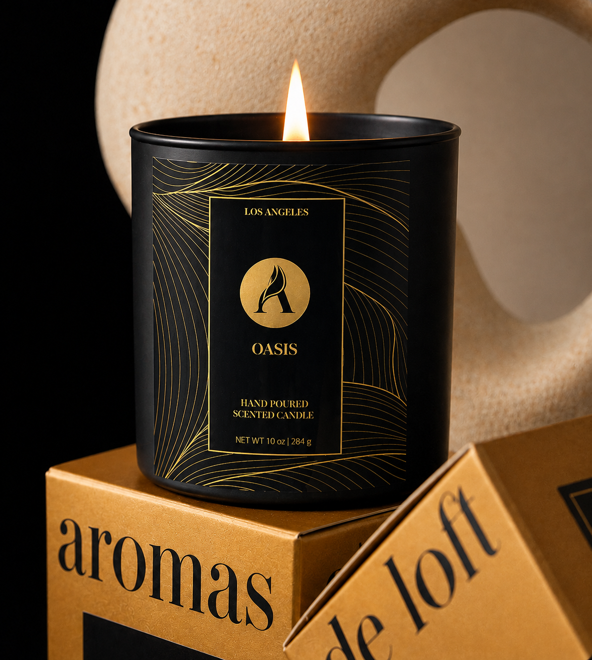 A black glass scented candle with a gold label reading 'OASIS' and 'Los Angeles' is lit and displayed on a gold box. The background features a beige abstract wall sculpture against a black backdrop.